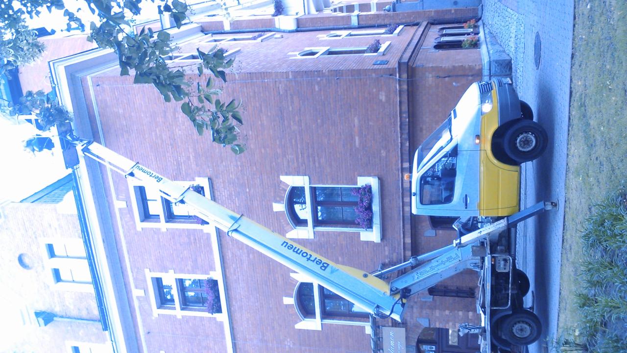 Żółto-biały podnośnik koszowy firmy Bertomeu na tle ceglanego budynku z oknami ozdobionymi kwiatami. Widok z dołu, obrócony o 90 stopni.