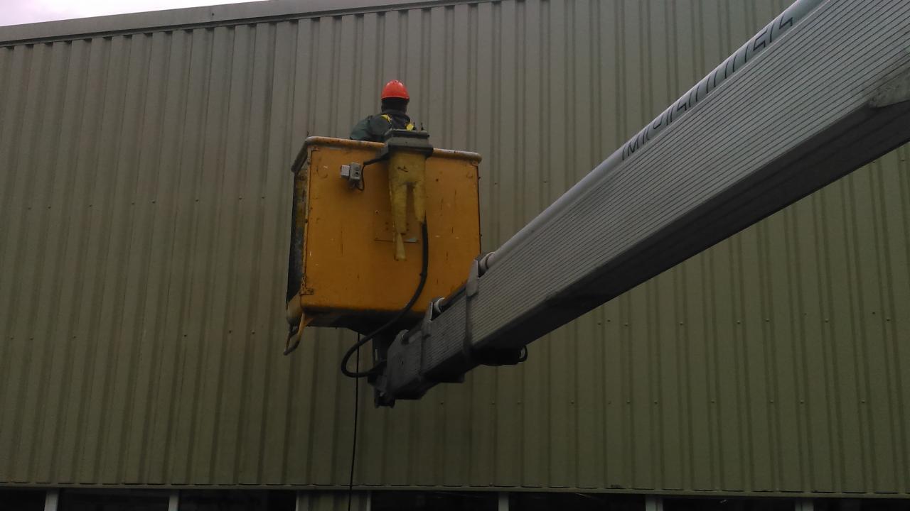 Żółty podnośnik koszowy z operatorem w kasku na tle blaszanej ściany budynku. Widoczny fragment wysięgnika z oznaczeniami.