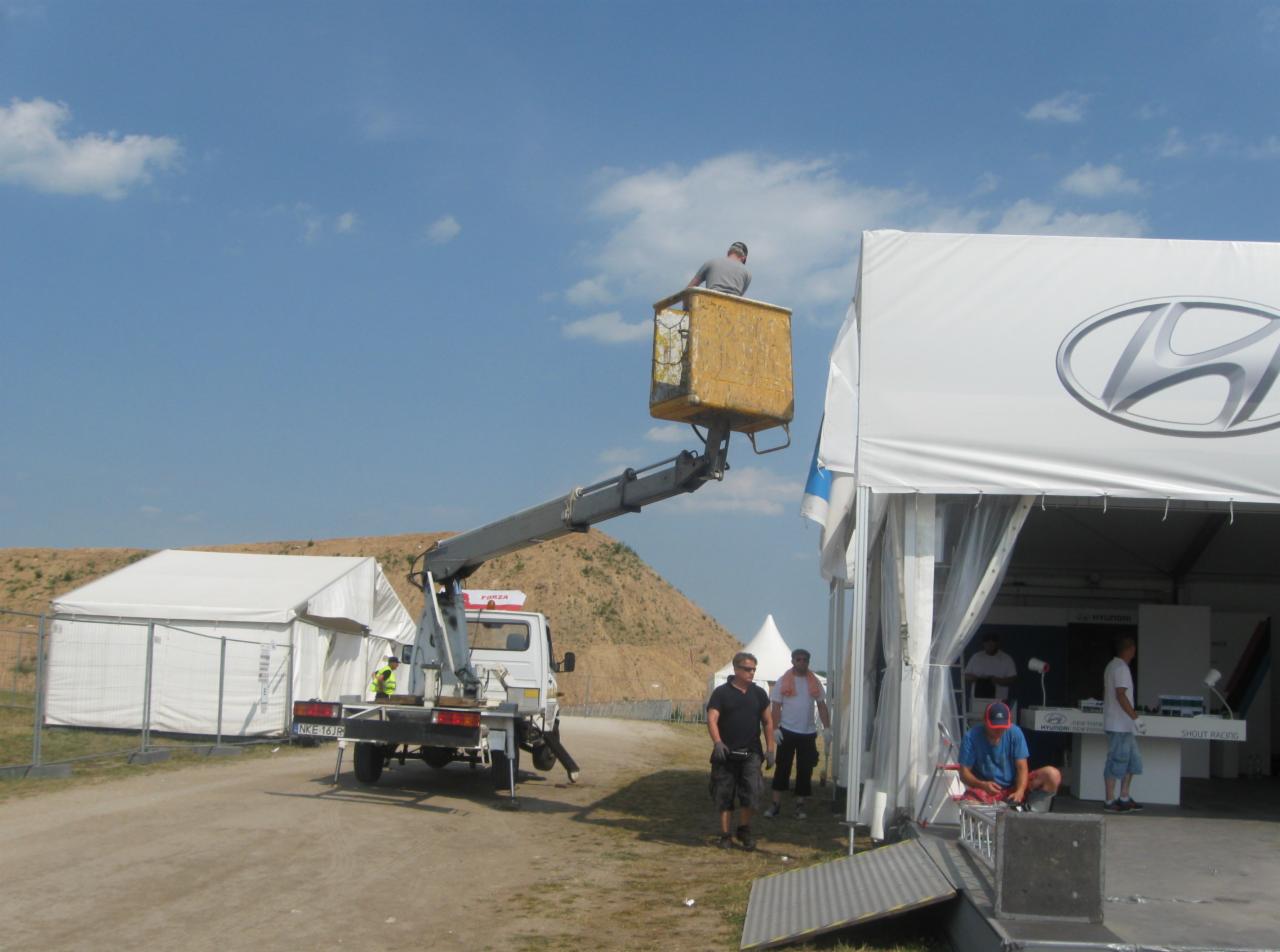 Pracownik w koszu podnośnika koszowego, montuje logo Hyundai na białym namiocie eventowym. W tle drugi namiot i wzgórze.