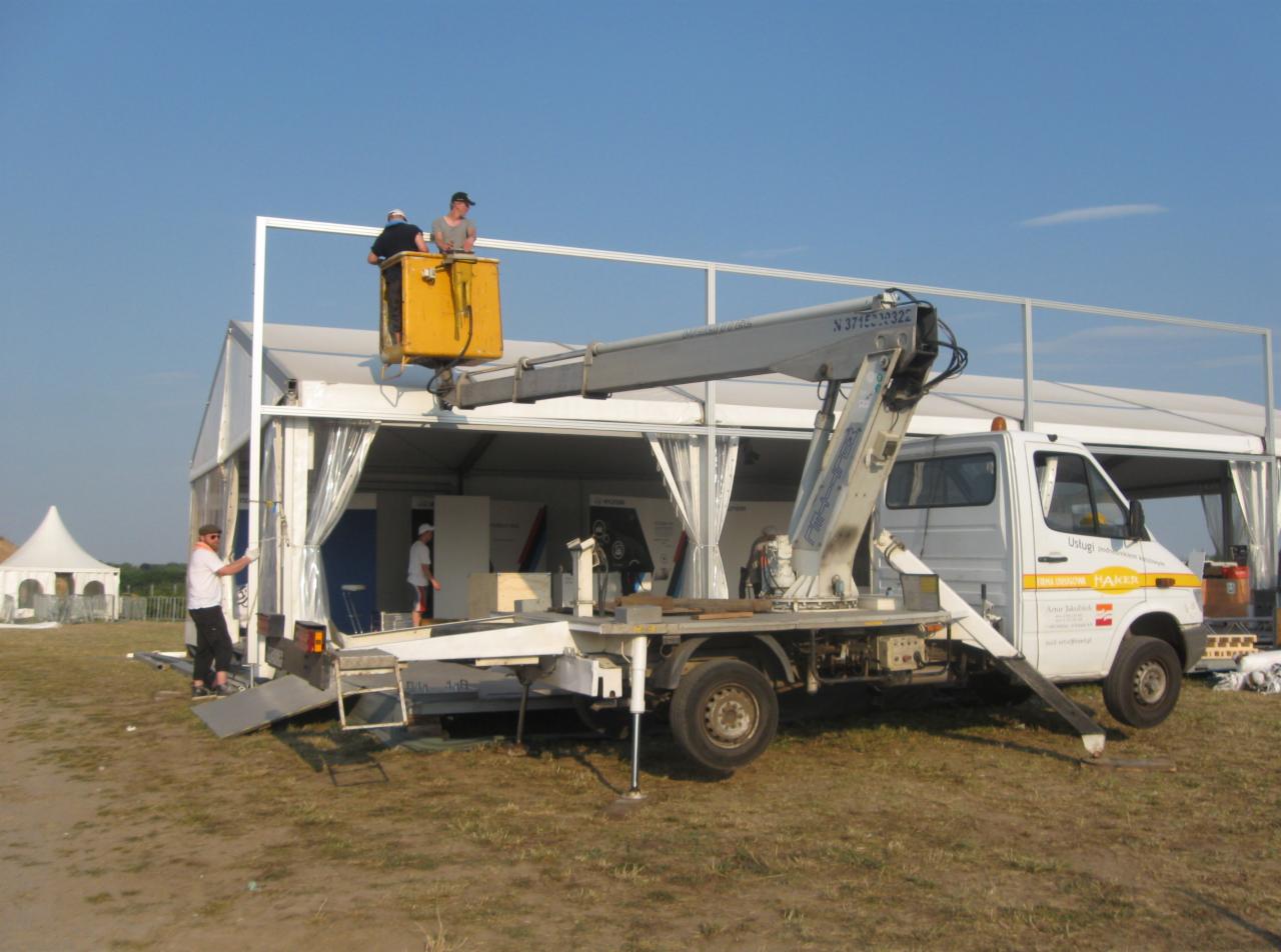 Montaż konstrukcji namiotowej z użyciem podnośnika koszowego; dwóch pracowników w żółtym koszu na tle białego namiotu i niebieskiego nieba.