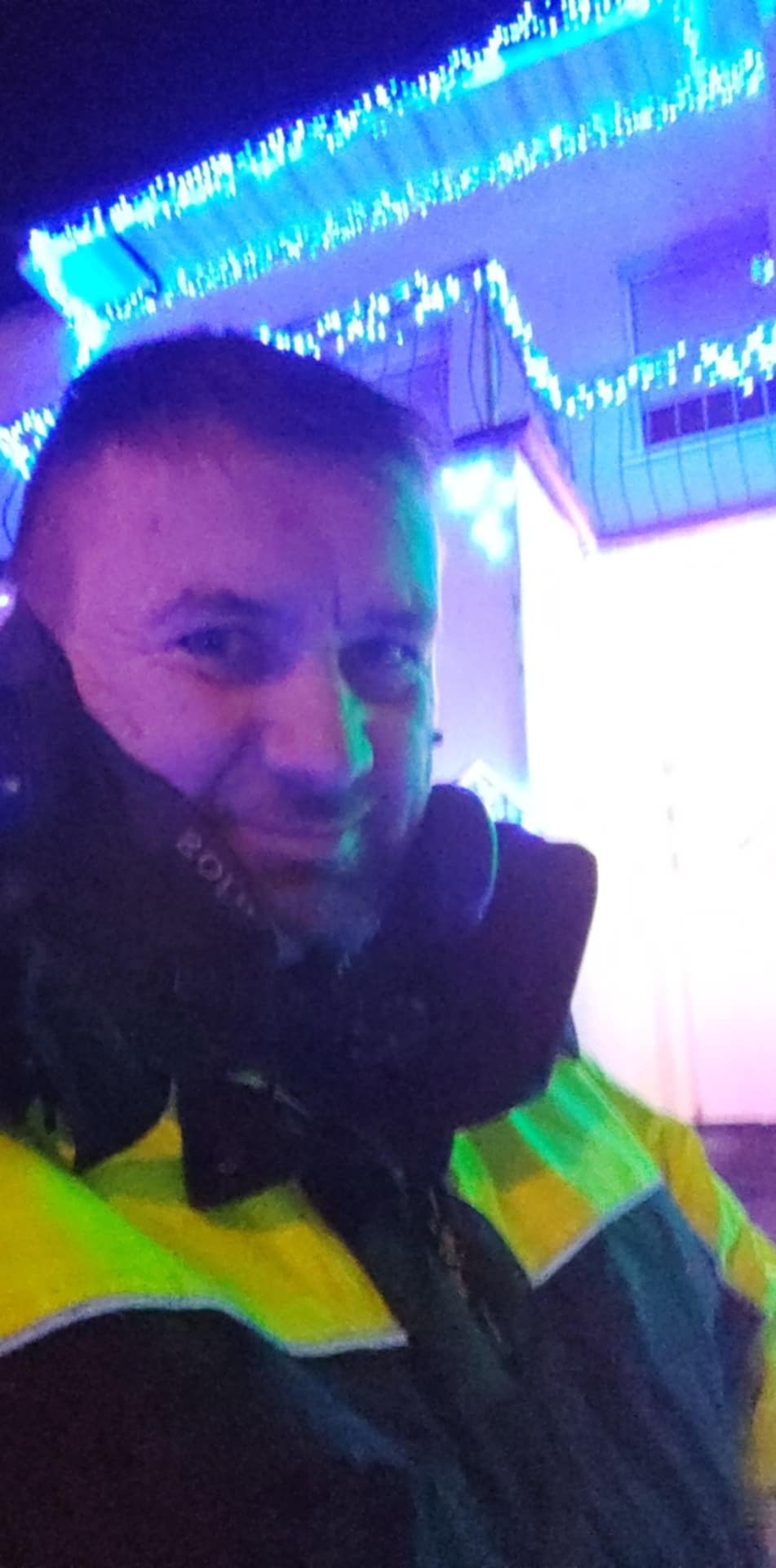 Uśmiechnięty mężczyzna w odblaskowej kurtce na tle budynku udekorowanego niebieskimi światełkami. Nocne ujęcie.