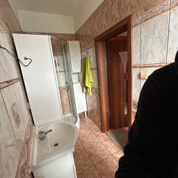 BorBud - Ukończona łazienka z kabiną prysznicową, umywalką z szafką i brązowymi płytkami. Widoczny fragment osoby w czarnej bluzie.