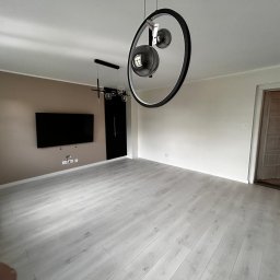 BorBud - Jasne panele podłogowe w przestronnym salonie z beżową ścianą, czarnym telewizorem i designerską lampą. Widoczne drzwi z osobą w środku.