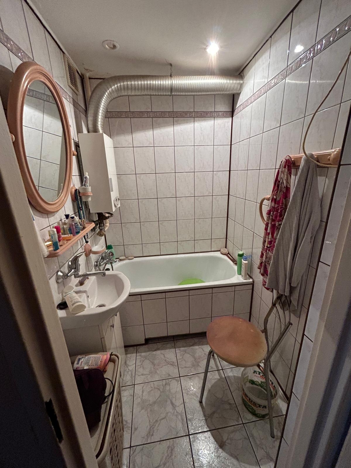 Wykończona łazienka z białą wanną, umywalką i grzejnikiem. Na ścianach białe płytki z dekoracyjnym paskiem. Widoczny okrągły lustro i metalowy przewód wentylacyjny.