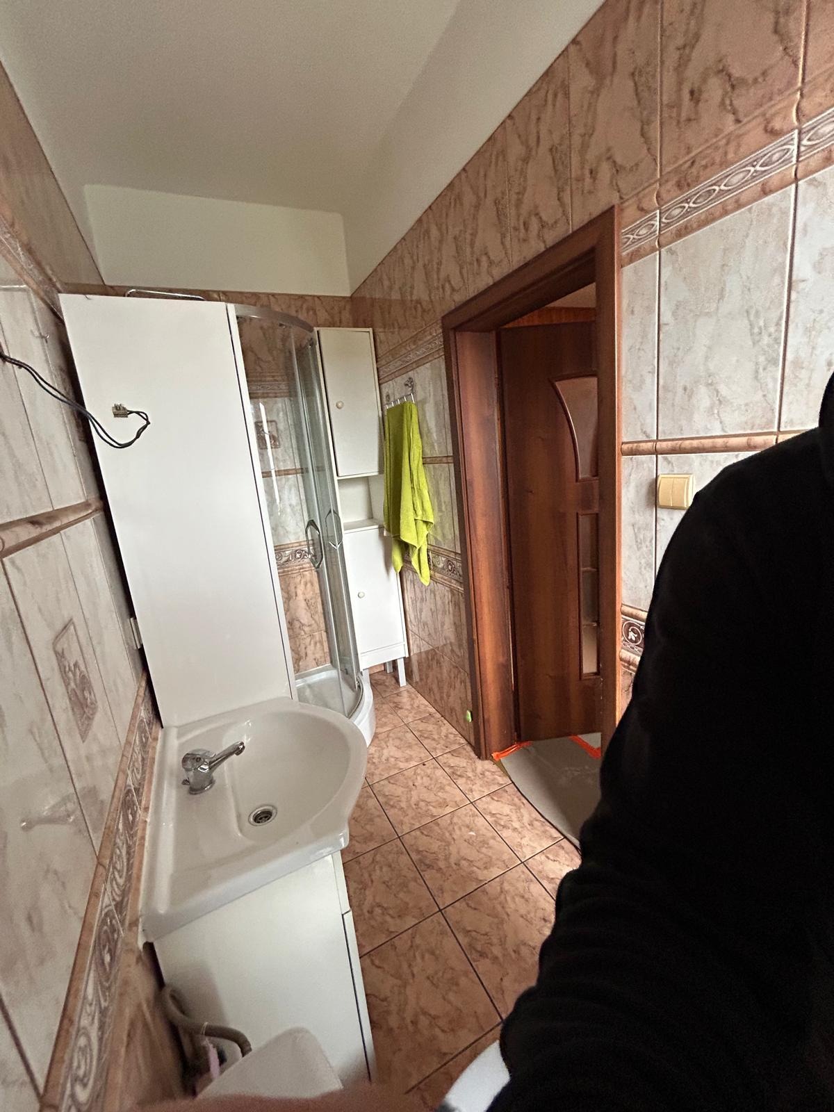 Ukończona łazienka z kabiną prysznicową, umywalką z szafką i brązowymi płytkami. Widoczny fragment osoby w czarnej bluzie.