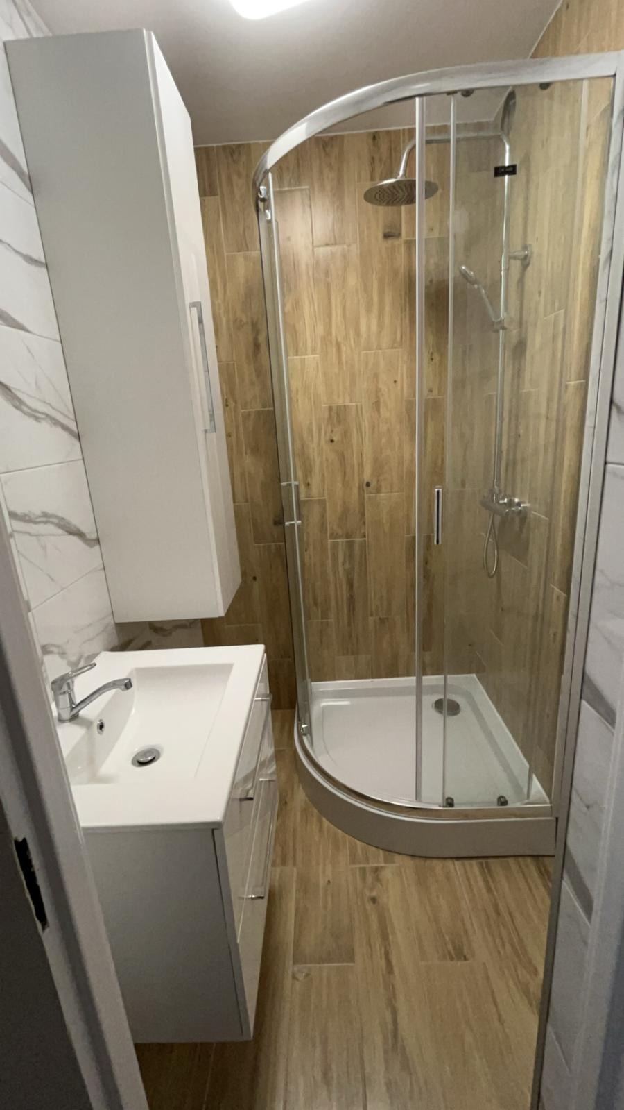 Nowoczesna łazienka po remoncie: kabina prysznicowa z zaokrąglonym szkłem, biała szafka z umywalką, płytki imitujące drewno na ścianach i podłodze. Minimalistyczny design.