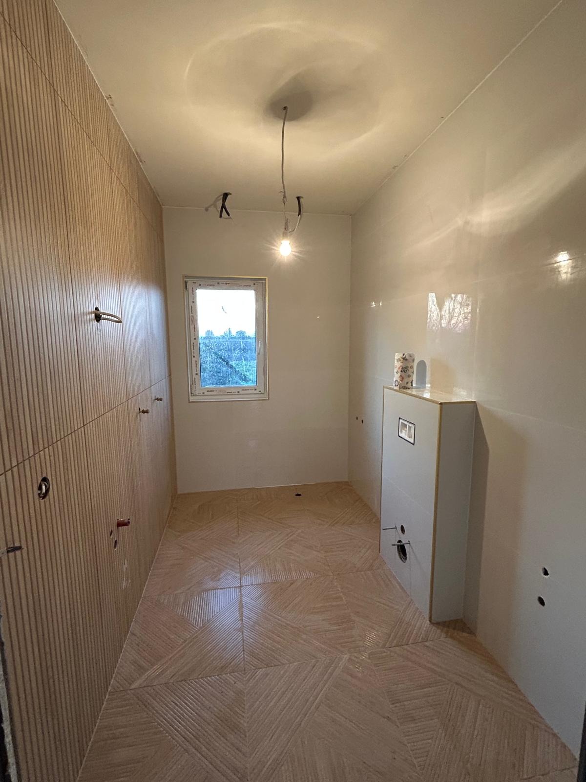 Wnętrze łazienki w trakcie wykańczania: ułożona podłoga z płytek w geometryczny wzór, częściowo wykończone ściany z dekoracyjnymi panelami i gładzią, wisząca lampa.