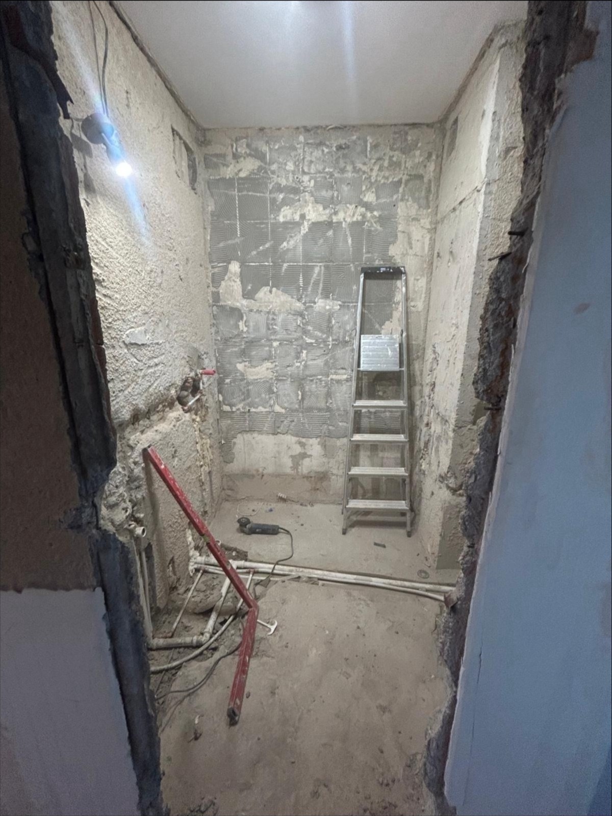 Łazienka w trakcie remontu: ściany z resztkami płytek, drabina, instalacja wodna, narzędzia. Widoczne ślady skuwania i przygotowania do dalszych prac.
