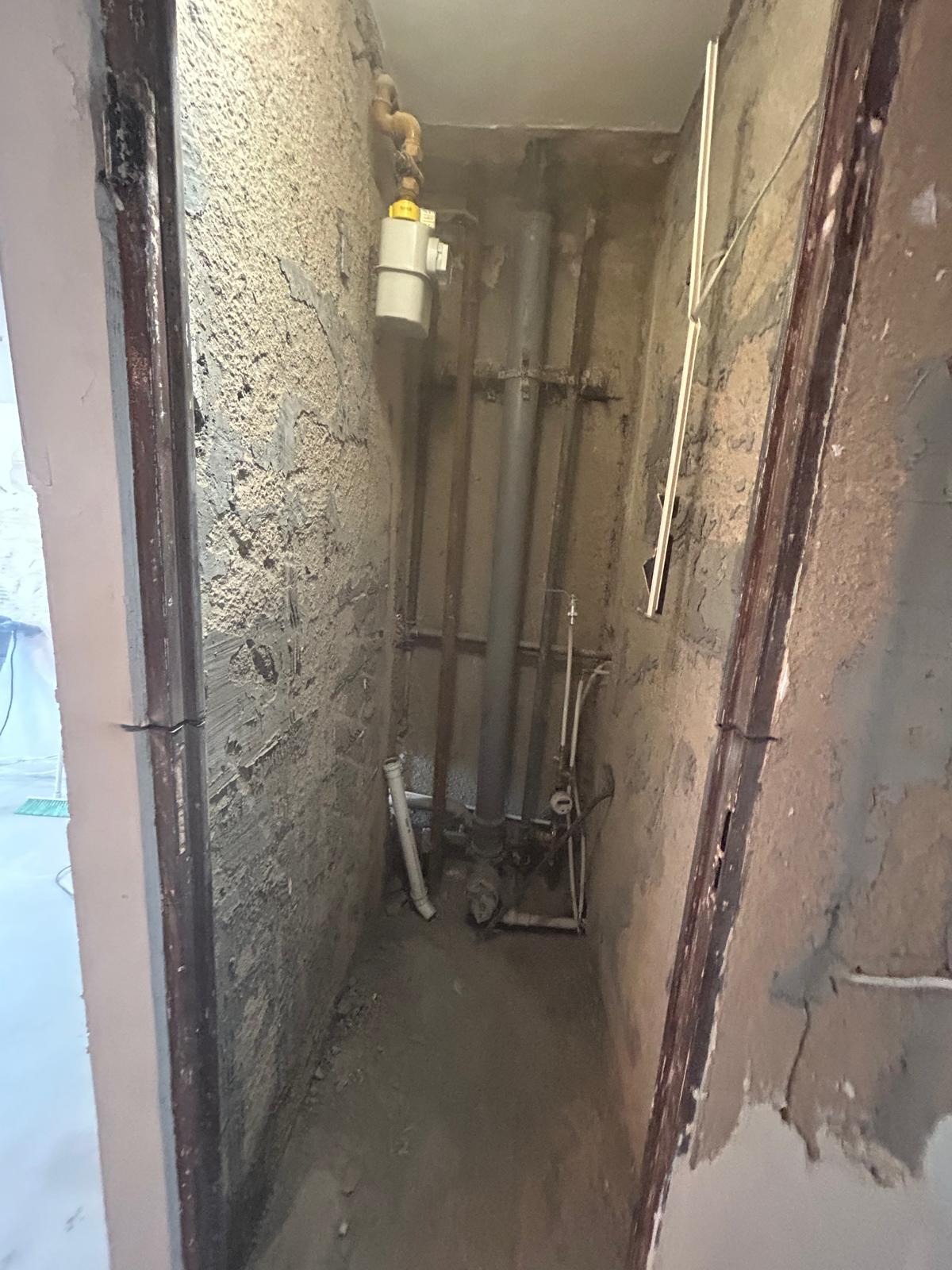 Wnęka z rurami i instalacją wodno-kanalizacyjną w trakcie remontu. Surowe ściany z widocznymi śladami prac murarskich. Fragment ościeżnicy drzwiowej po lewej stronie.