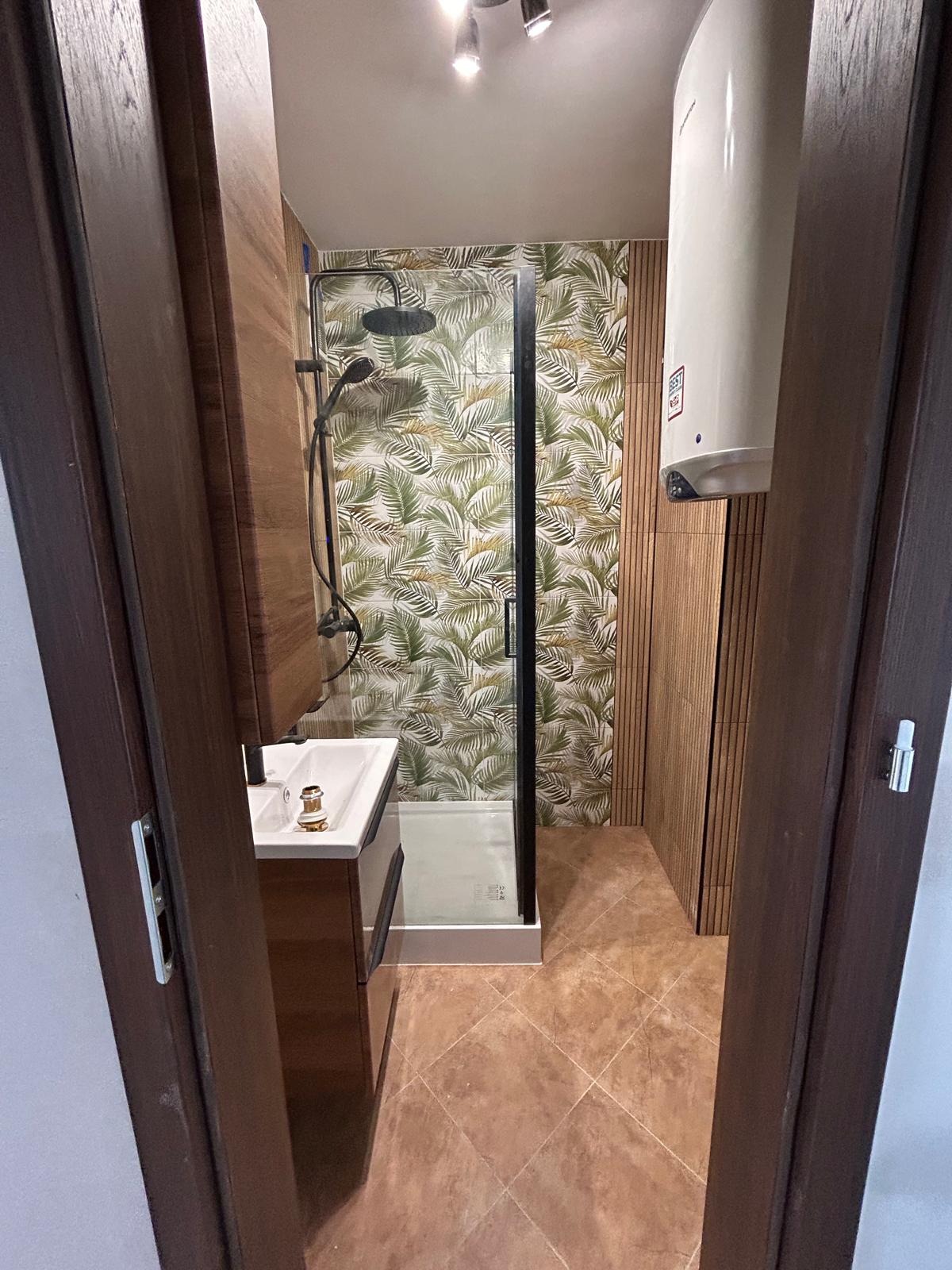Nowoczesna łazienka z kabiną prysznicową typu walk-in, umywalką z szafką w odcieniach brązu i płytkami imitującymi drewno oraz wzorzystą tapetą z motywem liści.