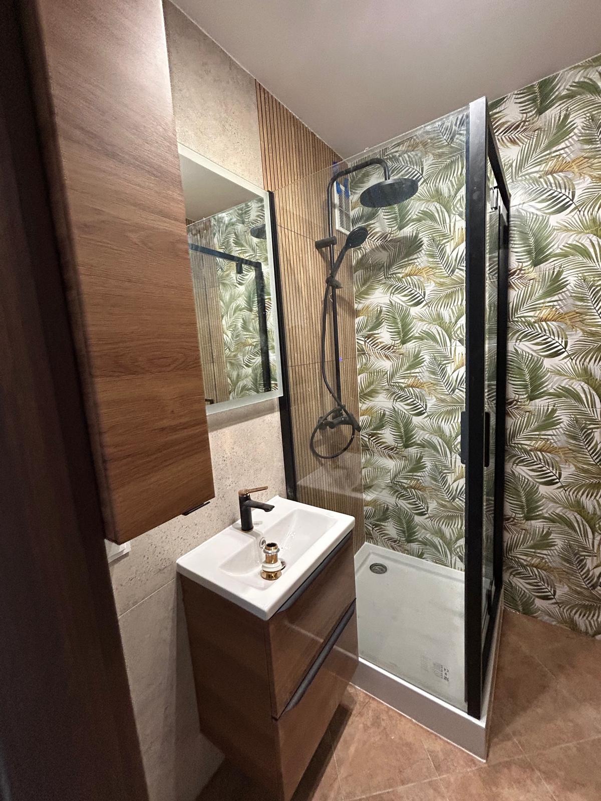 Nowoczesna łazienka z kabiną prysznicową typu walk-in, umywalką nablatową z szafką z drewna i lustrem. Ściana z motywem liści palmowych.