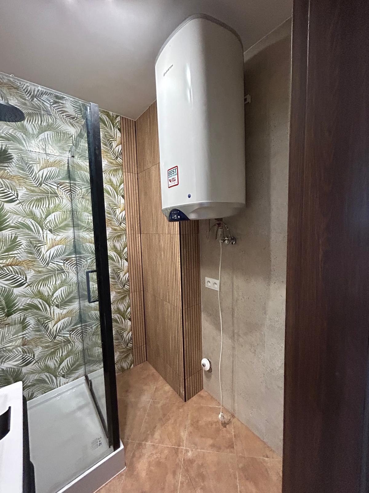 Biały bojler Ariston zamontowany w łazience z kabiną prysznicową i płytkami w liście palmy. Widoczne brązowe płytki podłogowe i drewnopodobne panele ścienne.