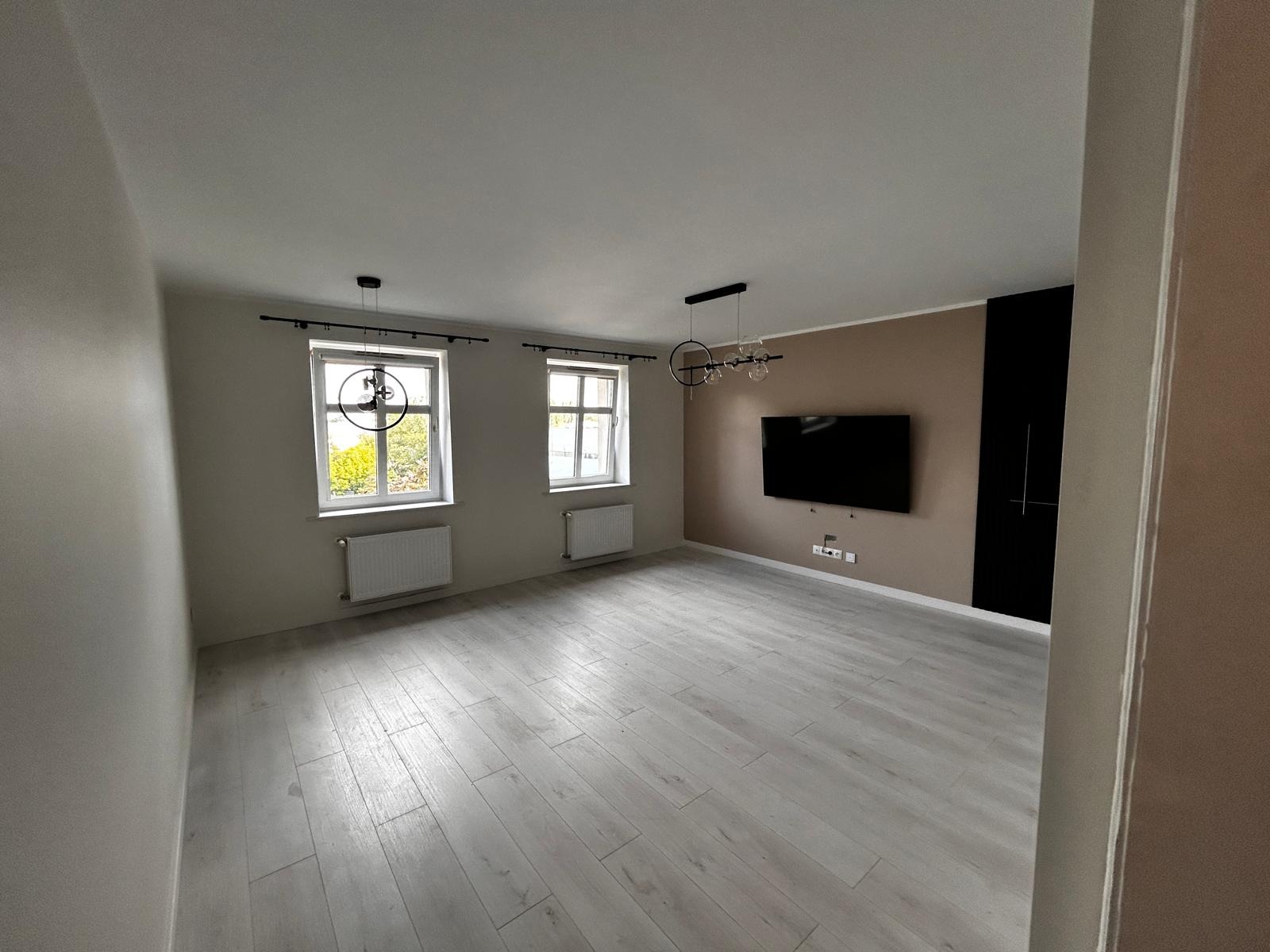 Jasne panele podłogowe w przestronnym pokoju z dwoma oknami, grzejnikami, telewizorem na ścianie i nowoczesnym oświetleniem. Ściany w odcieniach beżu i brązu.