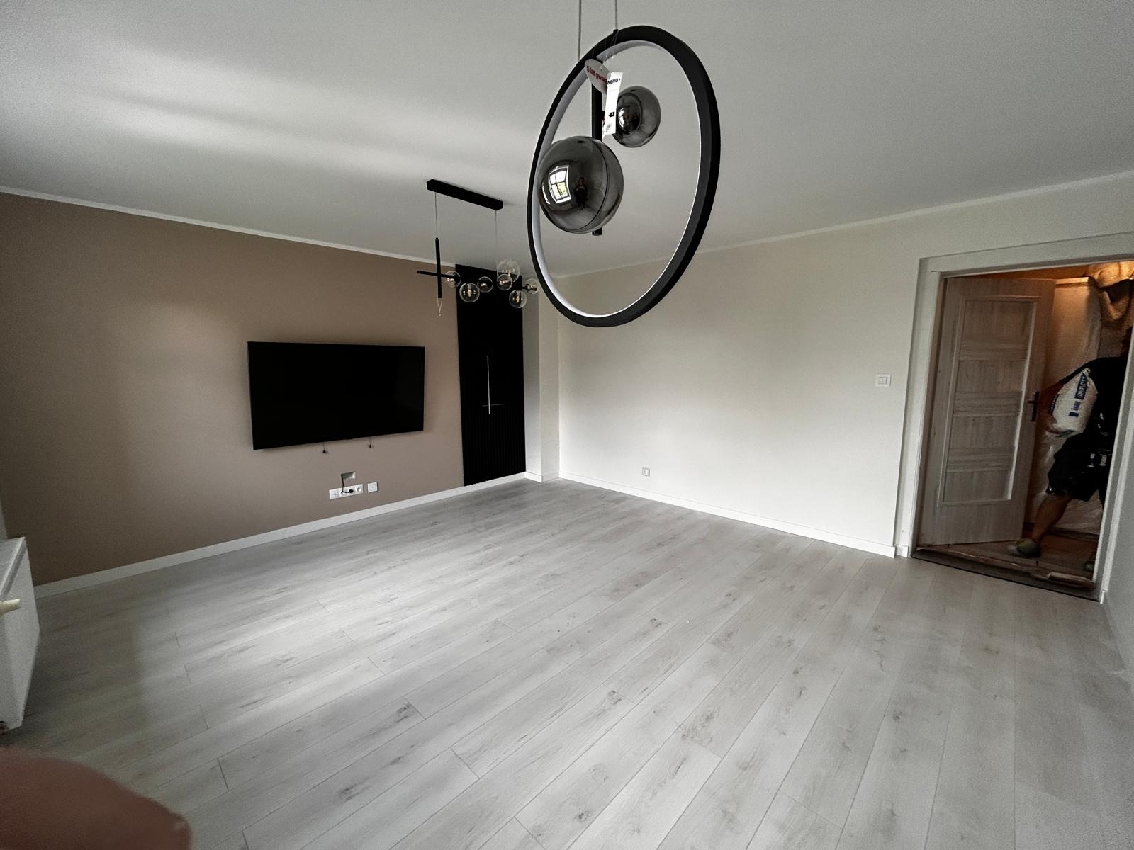 Jasne panele podłogowe w przestronnym salonie z beżową ścianą, czarnym telewizorem i designerską lampą. Widoczne drzwi z osobą w środku.