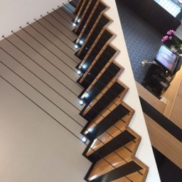 Piotr Konkol - Nowoczesne schody z drewnianymi stopniami i czarnymi elementami, oświetlone punktowo, z balustradą z linek, widziane z dołu pod kątem.
