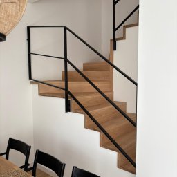 Usługi Stolarskie PIOTR KONKOL - Nowoczesne schody z jasnego drewna z czarną, minimalistyczną balustradą, widoczne z dołu, z fragmentem stołu i krzeseł w dolnej części kadru.