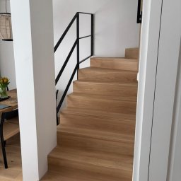 Usługi Stolarskie PIOTR KONKOL - Jasne, drewniane schody z czarną, metalową balustradą w nowoczesnym wnętrzu. Widok z dołu, z fragmentem stołu i krzesła w tle. Minimalistyczny design.