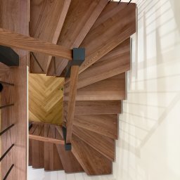 Usługi Stolarskie PIOTR KONKOL - Spiralne schody z drewna o ciepłym odcieniu, z czarnymi metalowymi balustradami, widziane z góry. Parkiet w jodełkę na podeście. Minimalistyczny design.