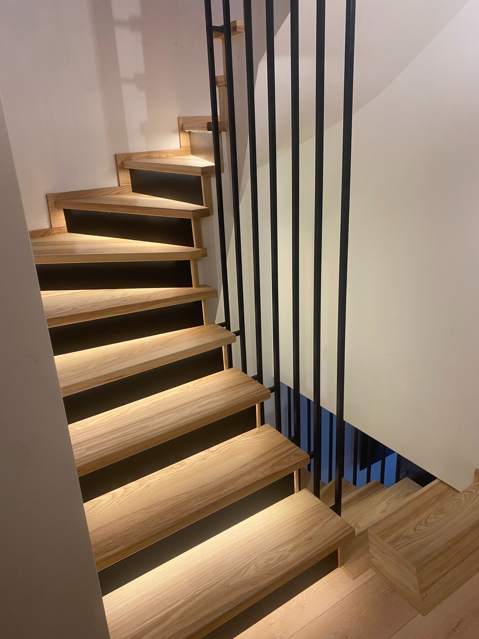 Spiralne schody z jasnego drewna z podświetleniem LED i czarną, minimalistyczną balustradą. Nowoczesny design, ciepłe oświetlenie.