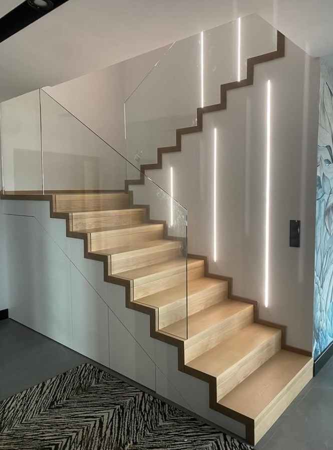 Nowoczesne schody z jasnego drewna z szklaną balustradą i subtelnym oświetleniem LED wzdłuż ściany, tworzące minimalistyczny design wnętrza.