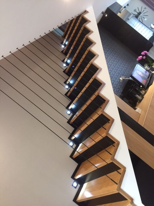 Nowoczesne schody z drewnianymi stopniami i czarnymi elementami, oświetlone punktowo, z balustradą z linek, widziane z dołu pod kątem.