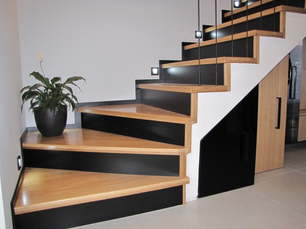 Nowoczesne schody z drewnianymi stopniami i czarnymi elementami, oświetleniem LED i rośliną doniczkową u podstawy. Minimalistyczny design z akcentem na naturalne materiały.