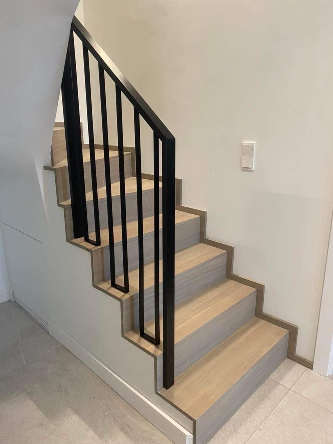 Jasne, drewniane schody z czarną, minimalistyczną balustradą w nowoczesnym wnętrzu. Widoczne detale wykończenia i subtelne oświetlenie.