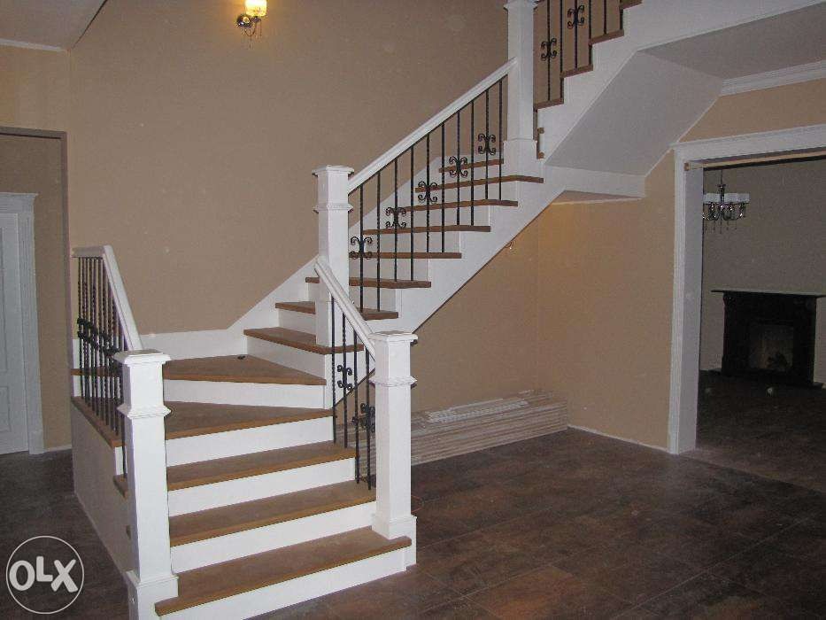 Białe schody drewniane z czarną, ozdobną balustradą w jasnym wnętrzu. Widoczne drewniane stopnie i fragment podłogi. W tle kominek i oświetlenie.