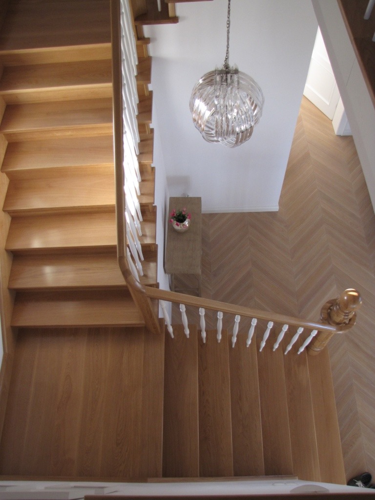 Jasne, drewniane schody z białą balustradą, widziane z góry, oświetlone przez nowoczesny żyrandol. Parkiet ułożony w jodełkę. Mała komoda z kwiatami przy ścianie.