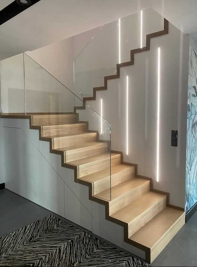 Nowoczesne schody z jasnego drewna z szklaną balustradą i subtelnym oświetleniem LED wzdłuż ściany. Minimalistyczny design z akcentem na naturalne materiały.