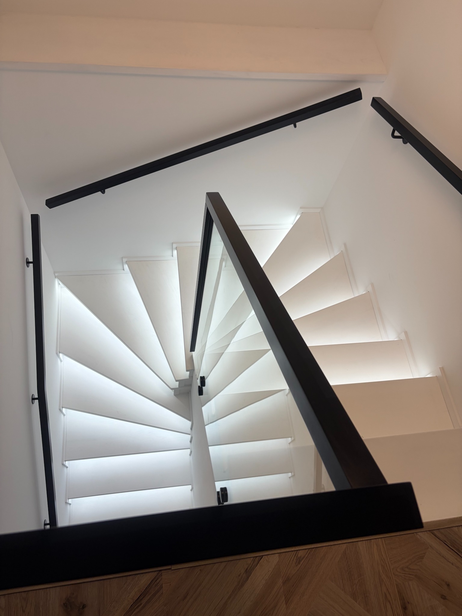 Spiralne schody z białymi stopniami, podświetleniem LED i czarną balustradą z elementami szkła, widziane z góry, z fragmentem drewnianej podłogi w dolnej części kadru.