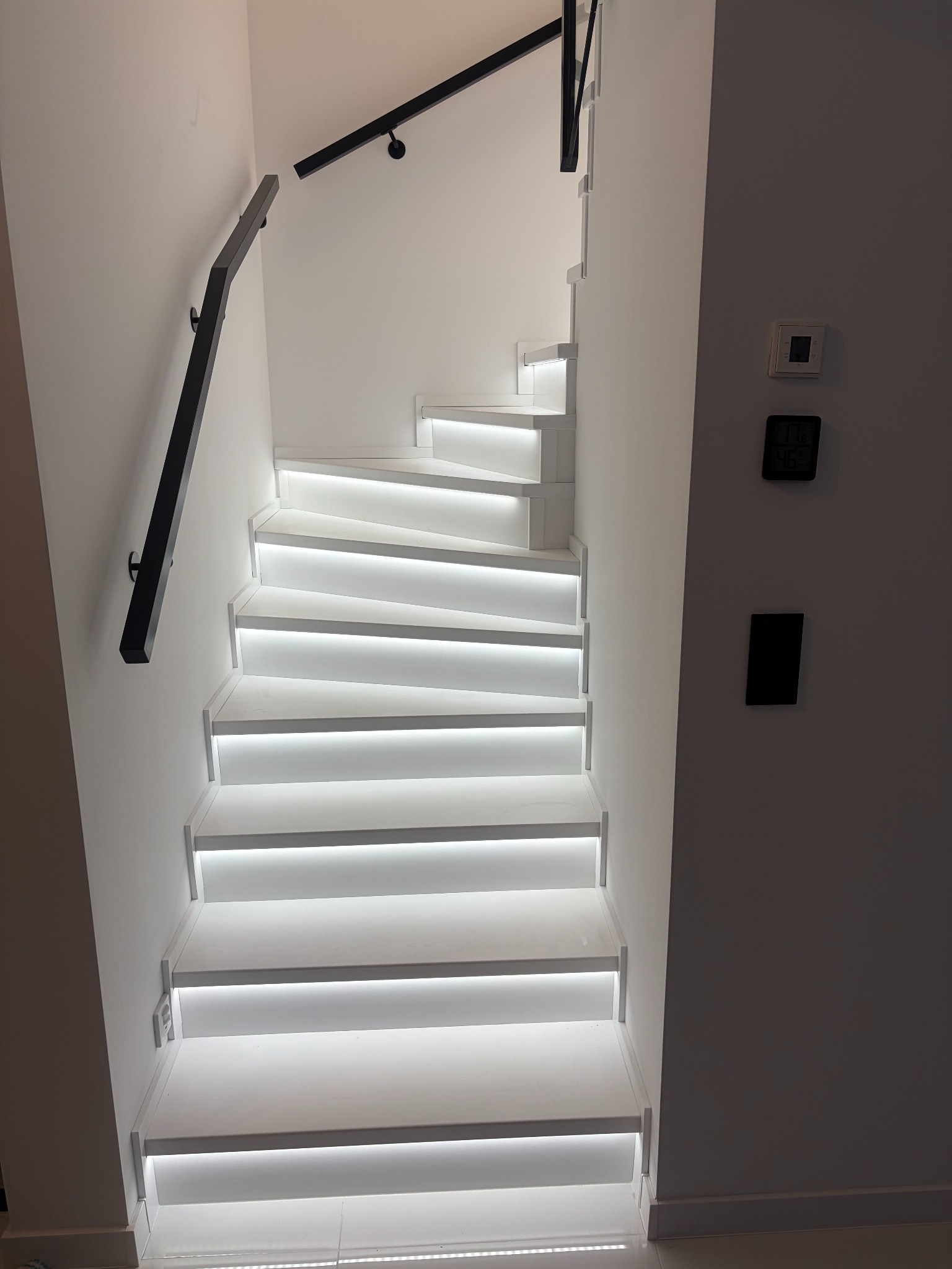 Nowoczesne, białe schody z podświetleniem LED i czarną balustradą, widok z dołu. Minimalistyczny design, jasne wnętrze.