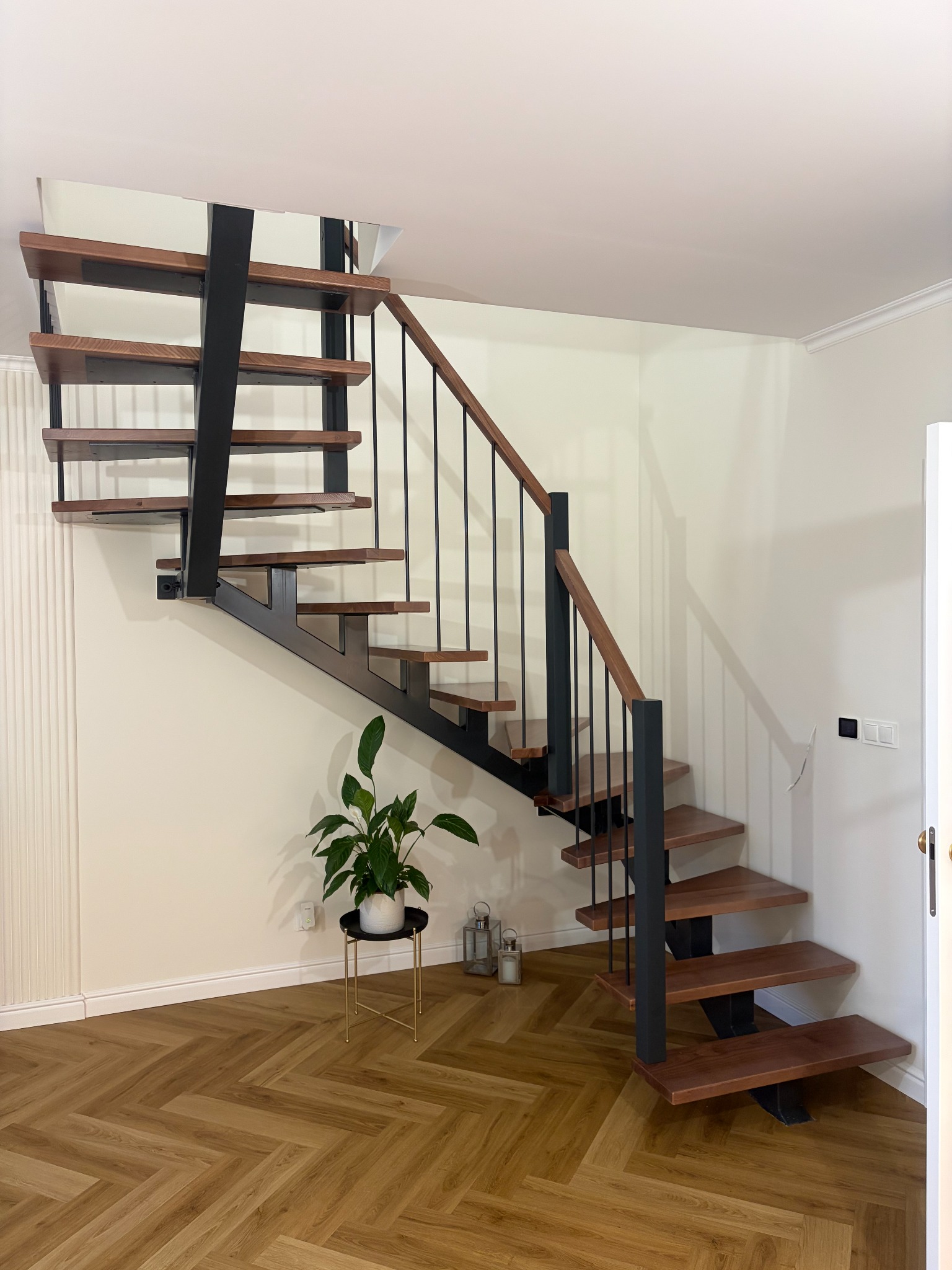 Nowoczesne schody z drewnianymi stopniami i czarną metalową konstrukcją, uzupełnione balustradą z drewnianym poręczą. W tle jasne ściany i podłoga w jodełkę.