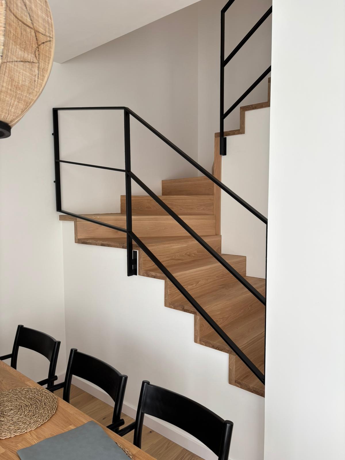 Nowoczesne schody z jasnego drewna z czarną, minimalistyczną balustradą, widoczne z dołu, z fragmentem stołu i krzeseł w dolnej części kadru.