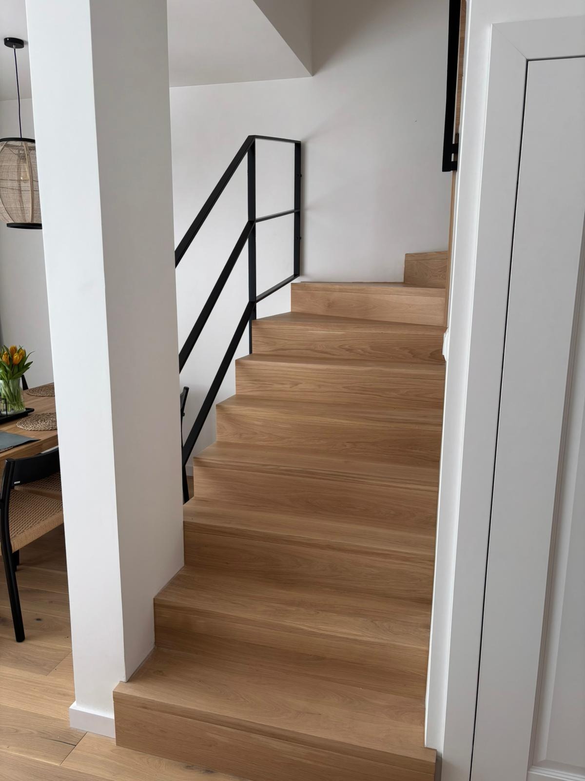 Jasne, drewniane schody z czarną, metalową balustradą w nowoczesnym wnętrzu. Widok z dołu, z fragmentem stołu i krzesła w tle. Minimalistyczny design.