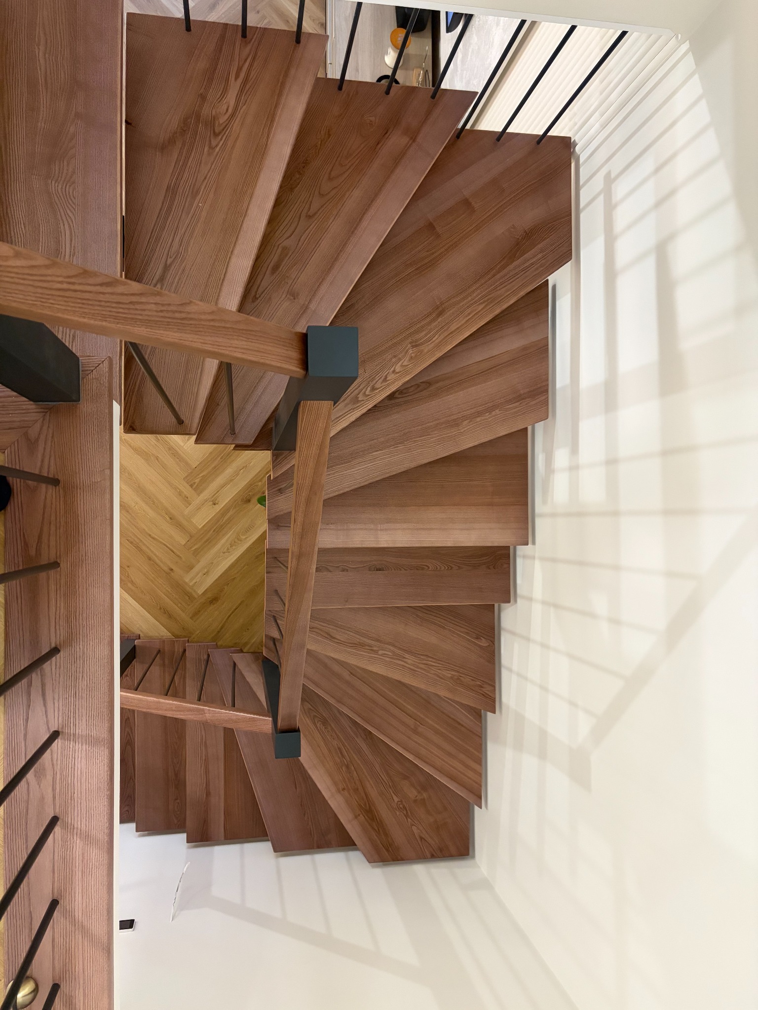 Spiralne schody z drewna o ciepłym odcieniu, z czarnymi metalowymi balustradami, widziane z góry. Parkiet w jodełkę na podeście. Minimalistyczny design.