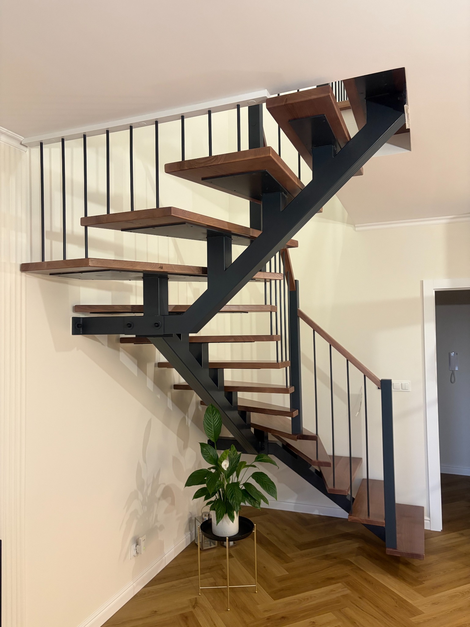 Nowoczesne schody z drewnianymi stopniami i czarną metalową konstrukcją, uzupełnione rośliną doniczkową na tle parkietu w jodełkę i jasnych ścian.