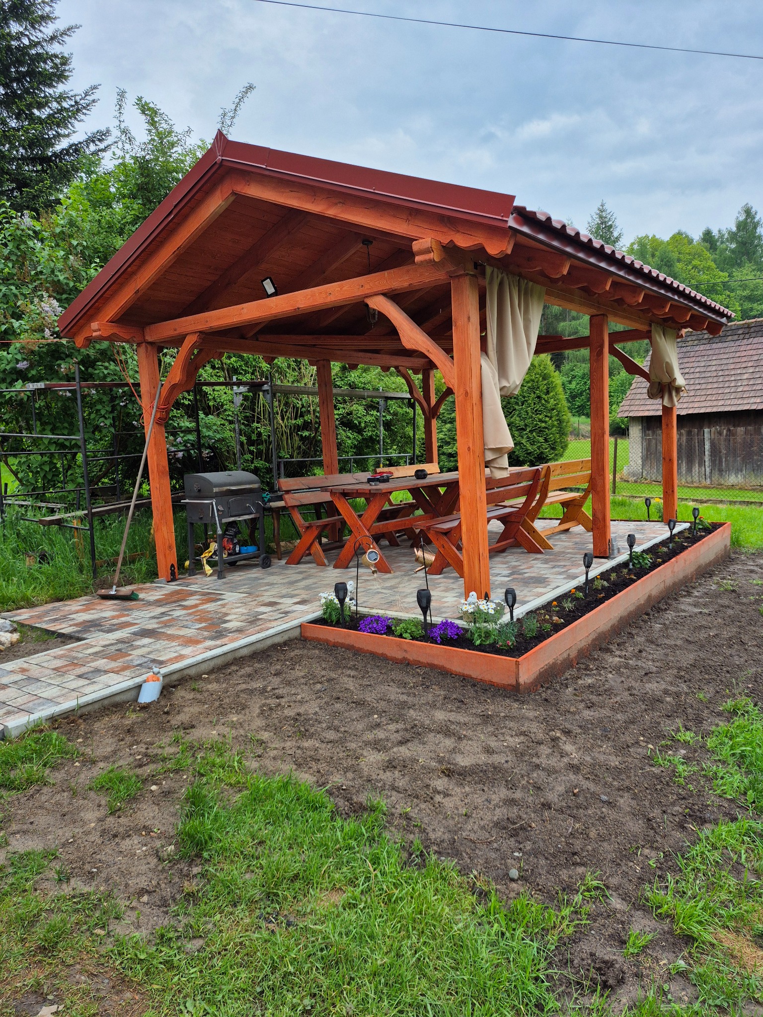 Drewniana altana z czerwonym dachem, stołem i ławkami, otoczona rabatą kwiatową i ścieżką z kostki, grill na zewnątrz. Widok na ogród i budynek w tle.