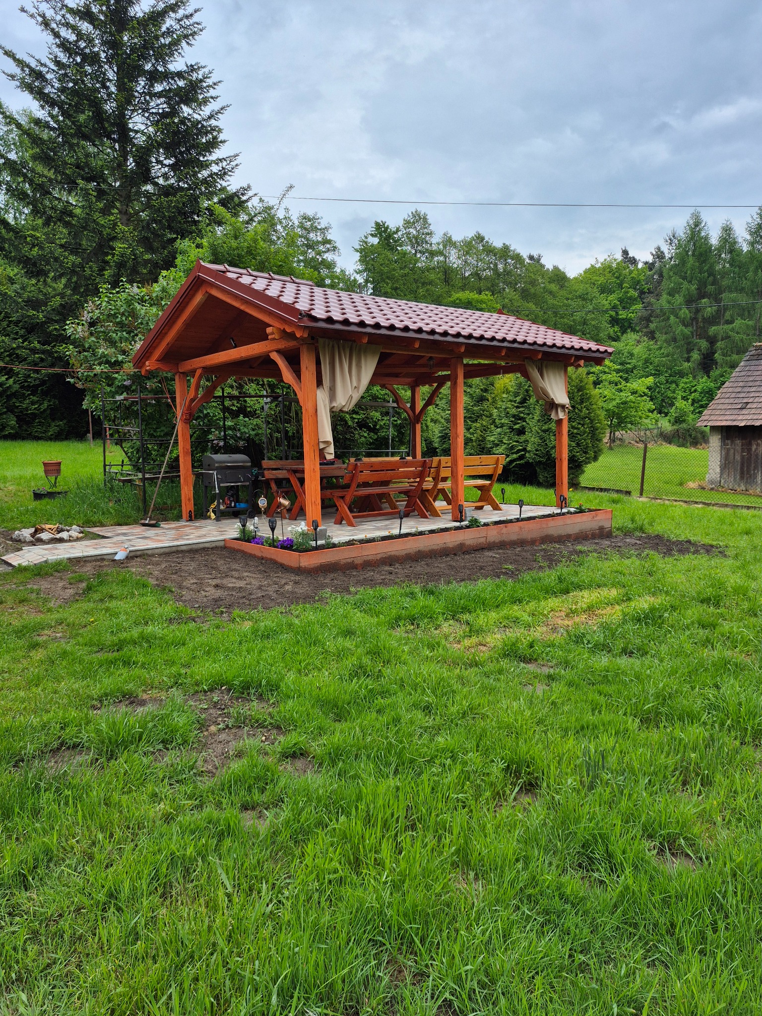 Drewniana altana z czerwonym dachem z blachodachówki, zasłonami i meblami ogrodowymi, otoczona trawnikiem i zielenią. Widok na las w tle.