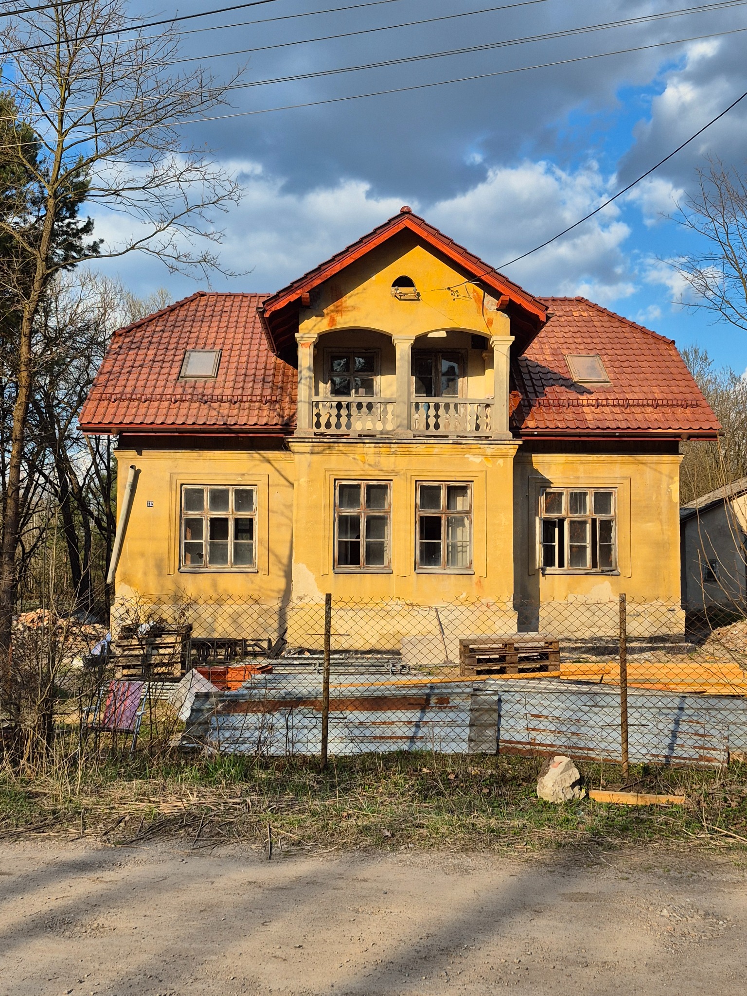 Żółty dom z balkonem i czerwoną dachówką, widoczny przez metalowe ogrodzenie. W tle błękitne niebo z chmurami i drzewa bez liści.