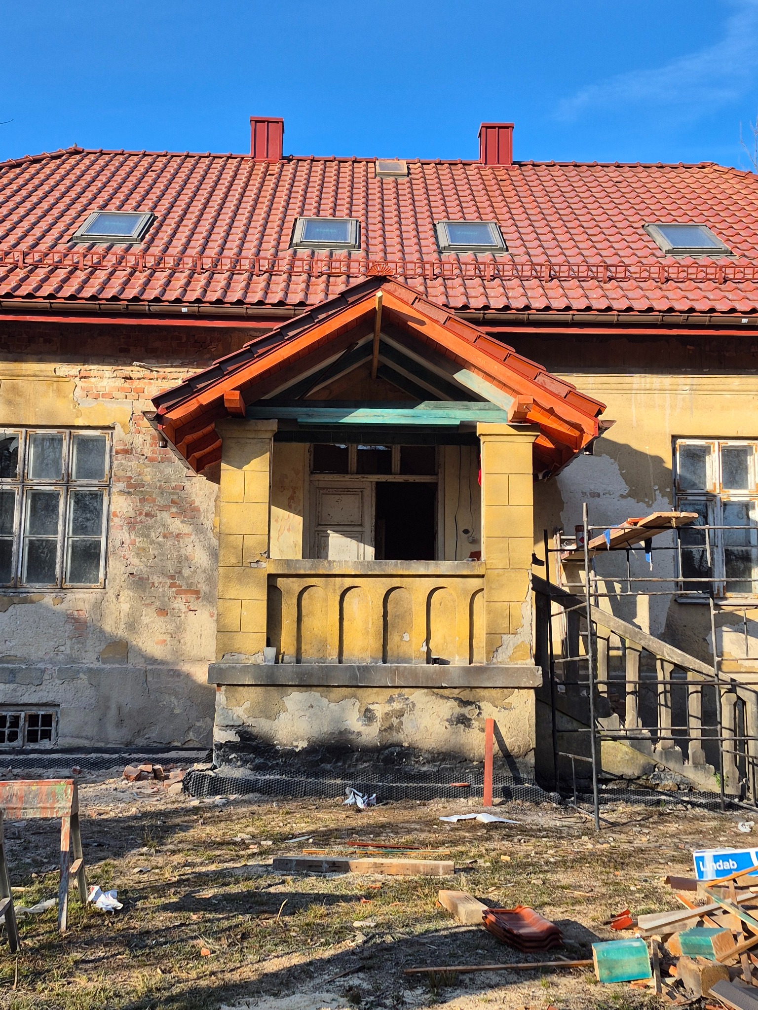 Ceglasty dach z lukarnami na budynku w trakcie renowacji. Widoczne rusztowania, elementy konstrukcyjne werandy i stare fragmenty elewacji. Słoneczny dzień.