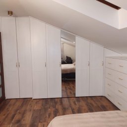 NOVALEX SPÓŁKA Z OGRANICZONĄ ODPOWIEDZIALNOŚCIĄ - Biała szafa na wymiar ze złotymi uchwytami, wkomponowana w skos dachu na poddaszu, z lustrzanym frontem i komodą, na drewnianej podłodze.