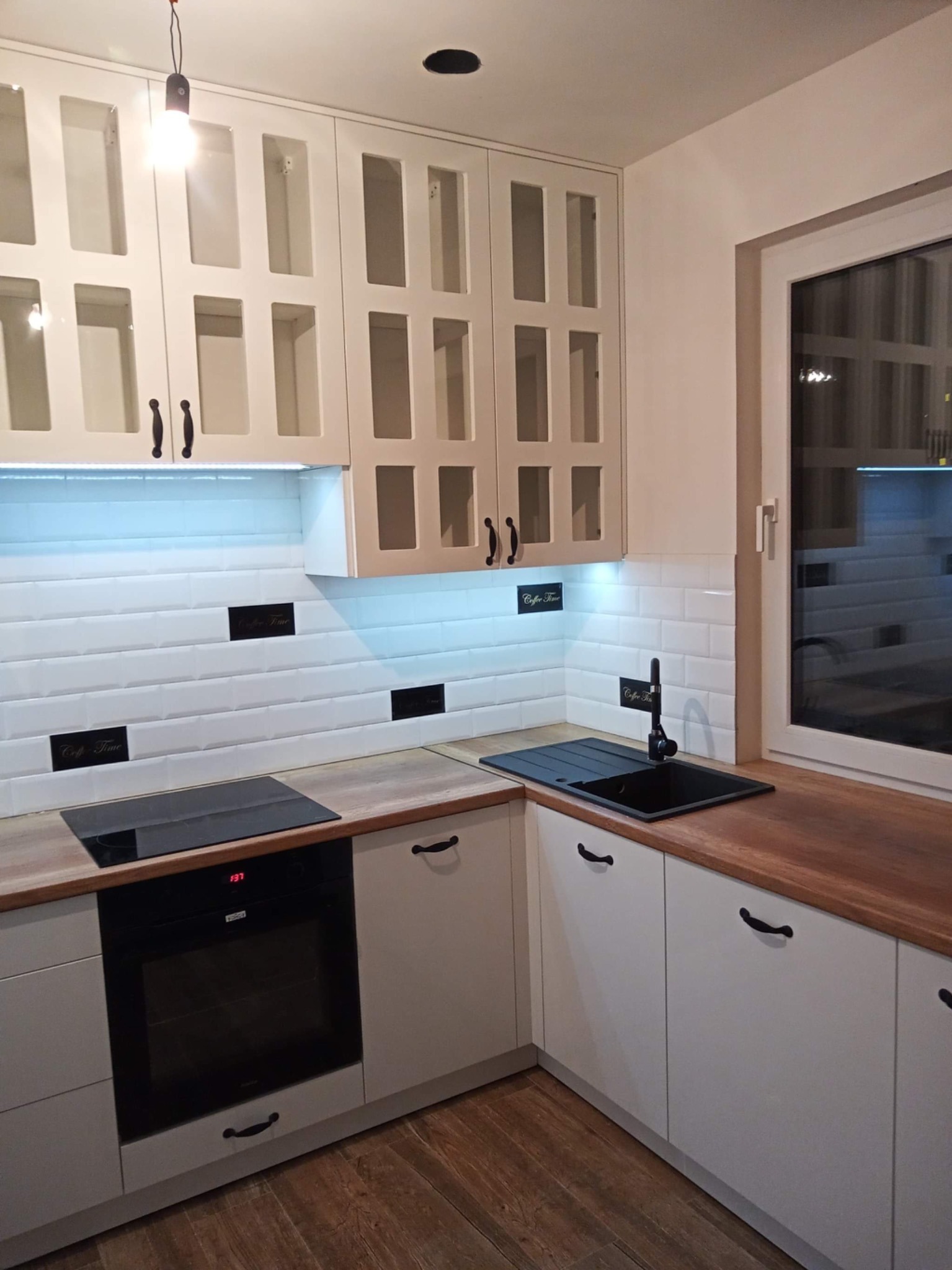 Biała kuchnia na wymiar z drewnianym blatem i czarnymi uchwytami, oświetlona ledami, z płytkami typu metro z dekoracyjnymi napisami. Widok na okno.