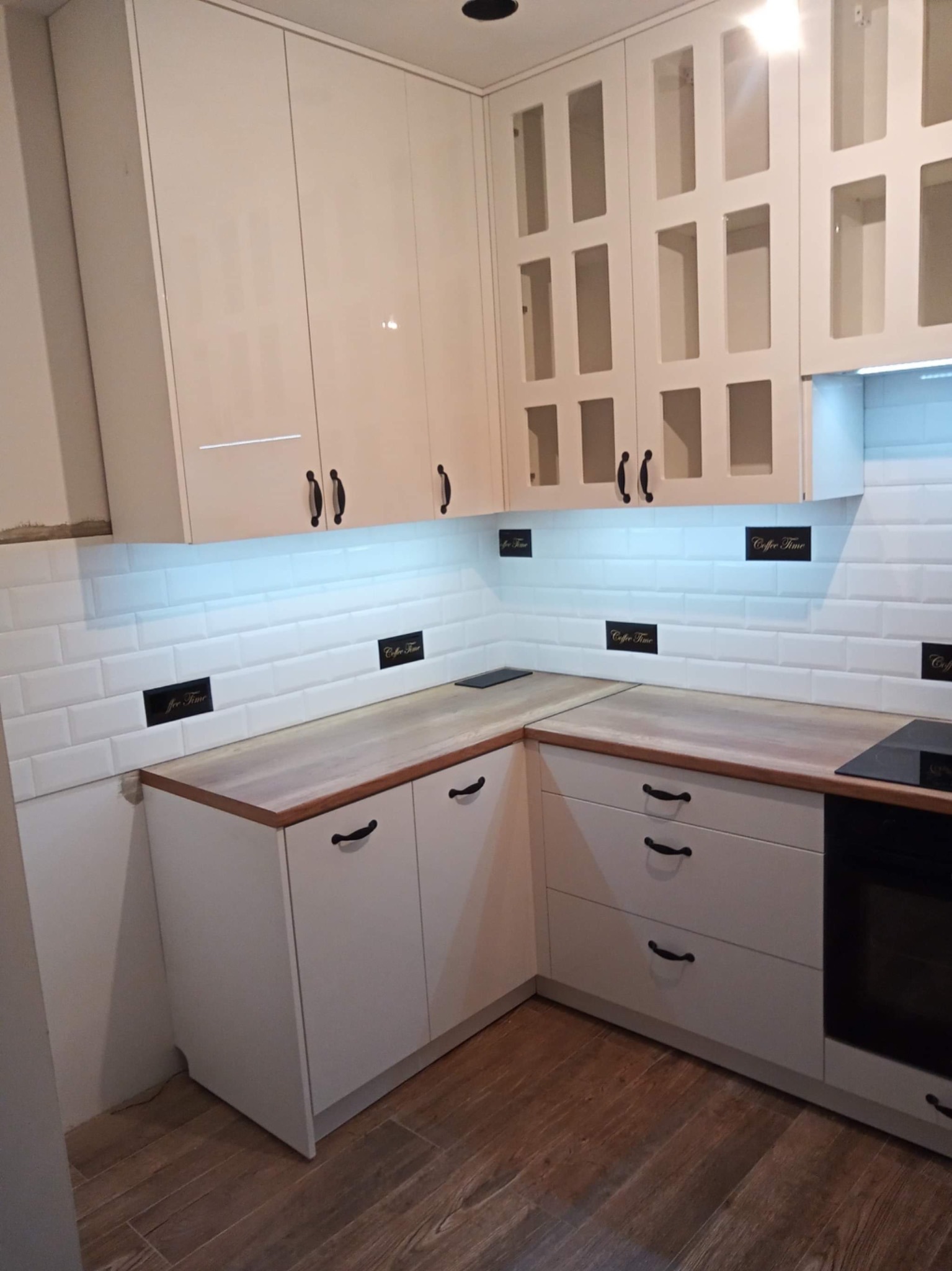 Jasna kuchnia z białymi, lakierowanymi frontami szafek, drewnianym blatem i czarnymi uchwytami. Na ścianie białe płytki typu metro z dekoracyjnymi wstawkami.