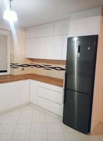 Jasna kuchnia z białymi szafkami i drewnianym blatem, uzupełniona o nowoczesną, srebrną lodówkę. Minimalistyczny design z akcentem na funkcjonalność i przestrzeń.