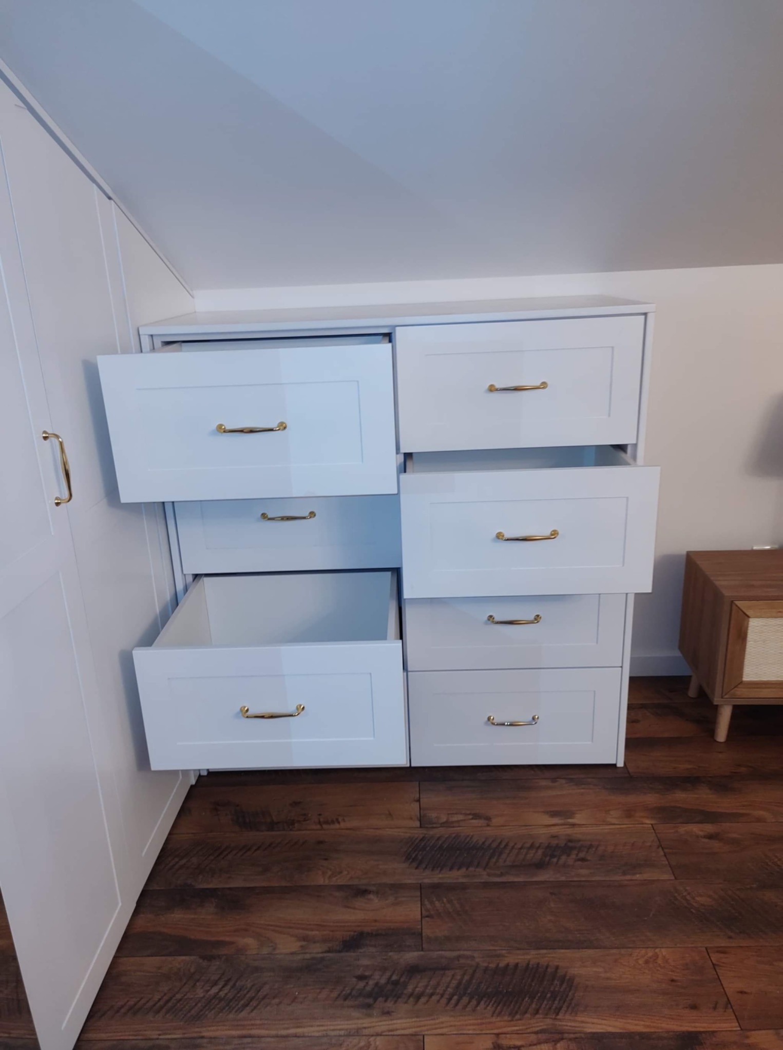 Biała szafa na wymiar z szufladami, część szuflad jest otwarta, widoczne złote uchwyty, drewniana podłoga. Szafa wkomponowana w skos dachu.