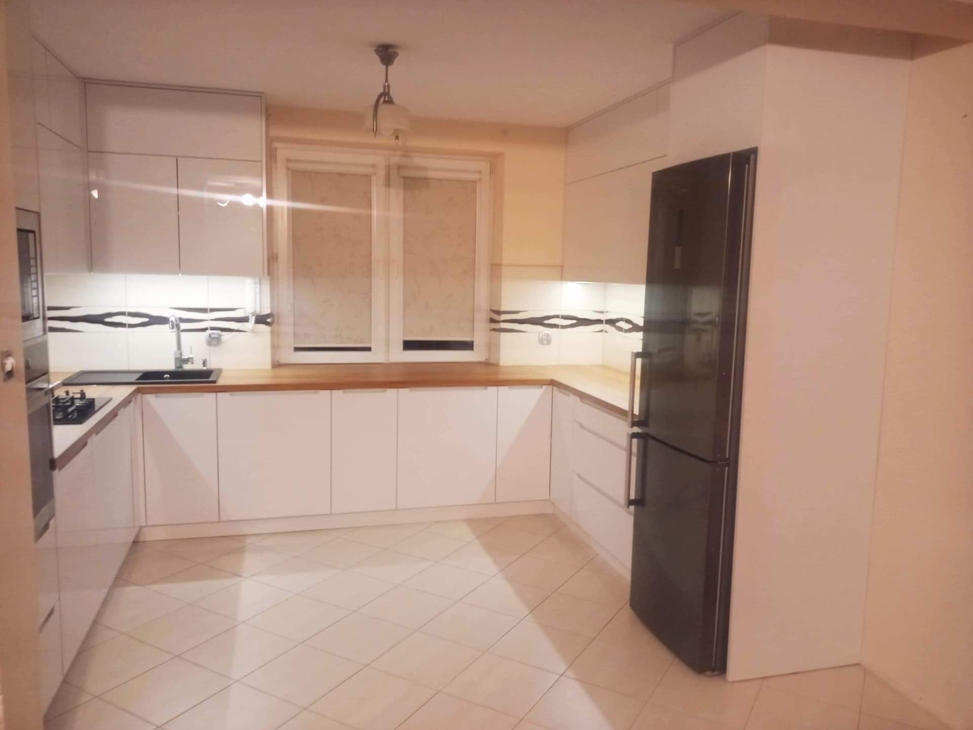 Jasna kuchnia z białymi szafkami, drewnianym blatem i czarnymi akcentami. Uwagę przykuwają płytki z motywem fal na ścianie nad blatem.