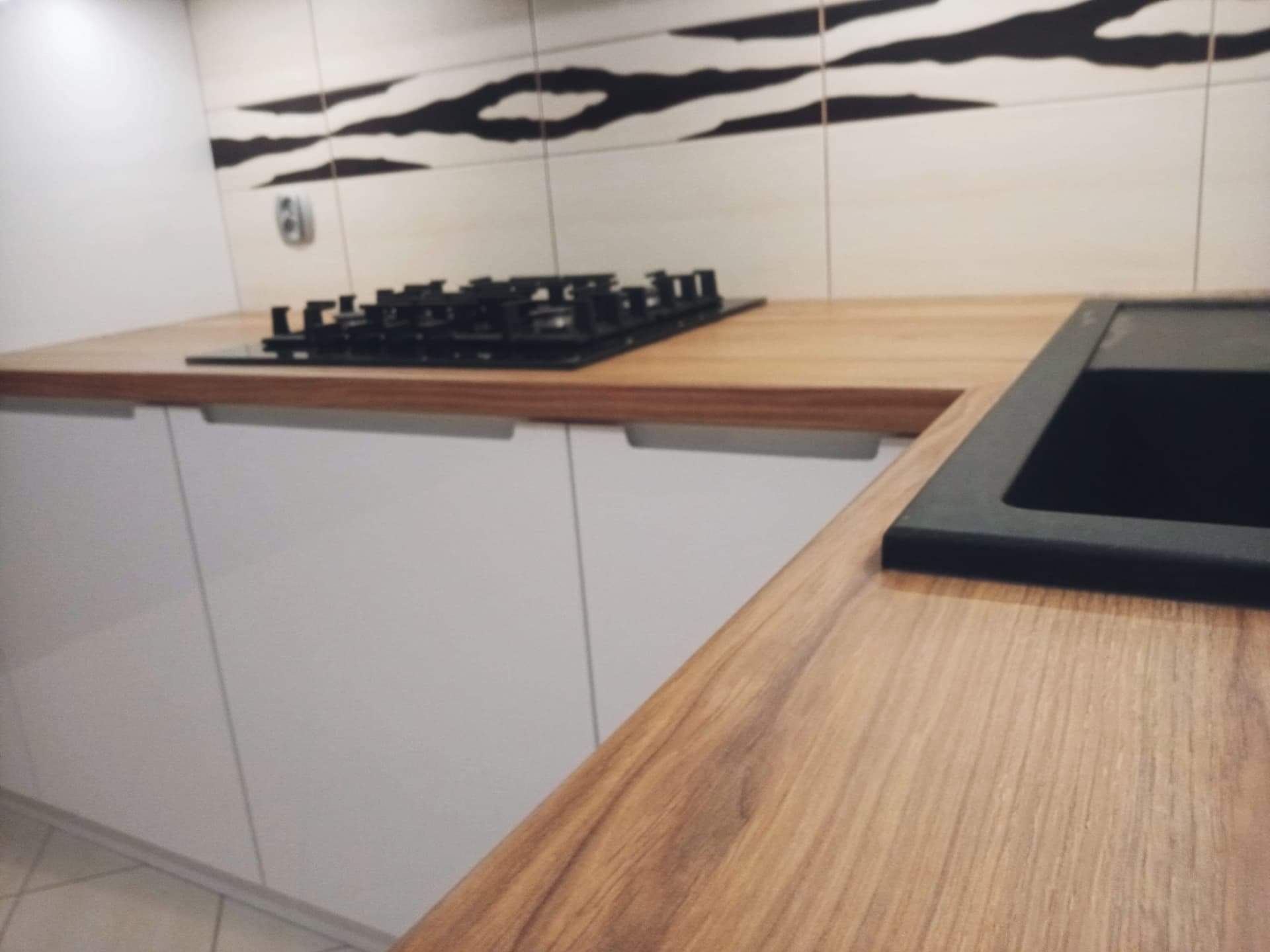 Jasny blat drewniany z czarną płytą grzewczą i zlewozmywakiem na tle białych szafek kuchennych i płytek z motywem zebry. Ujęcie z bliska, ukazujące detale wykończenia.