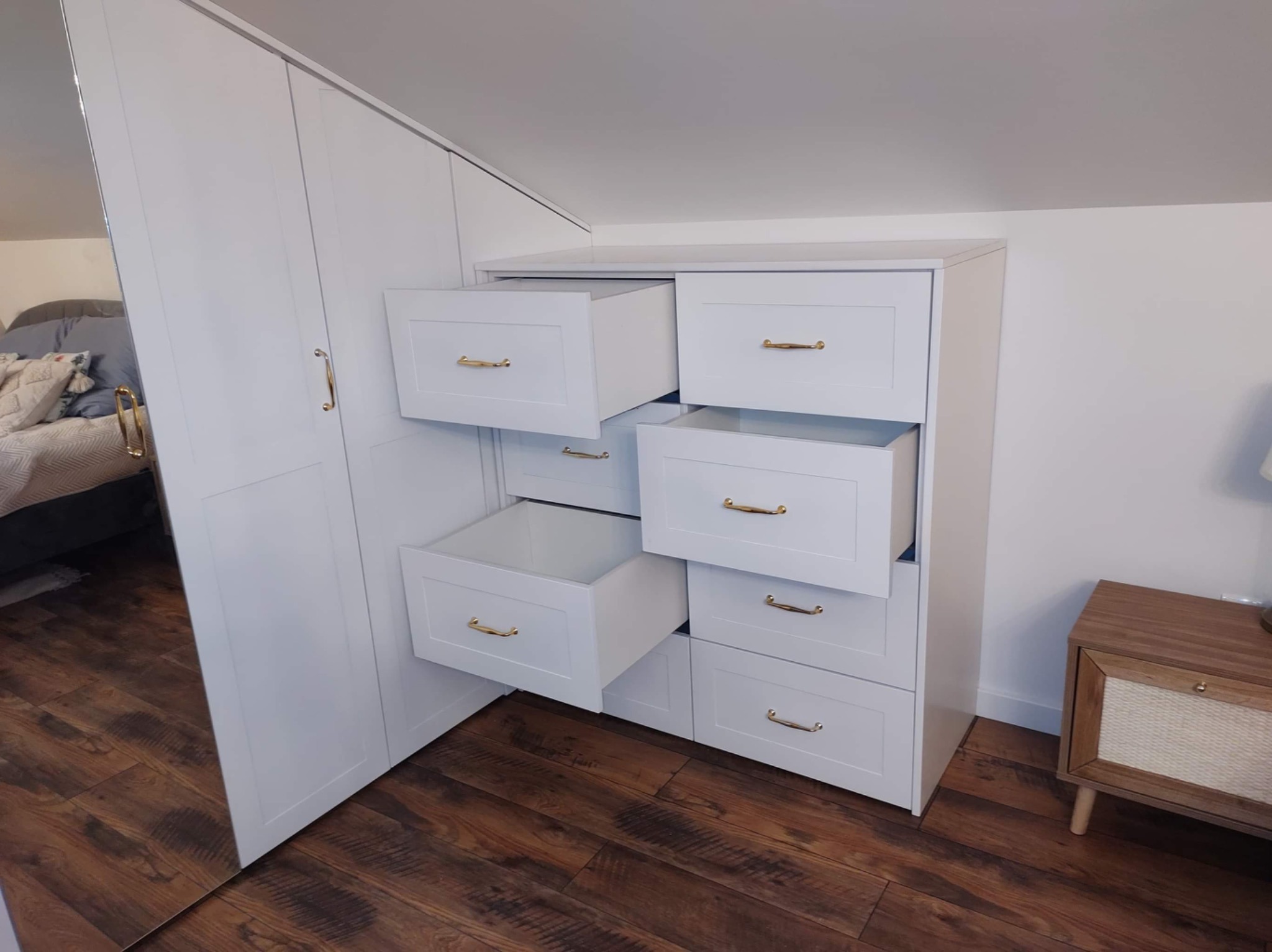 Biała szafa na wymiar z szufladami i złotymi uchwytami, częściowo otwarta, wkomponowana w skos pomieszczenia, z lustrzanym frontem.