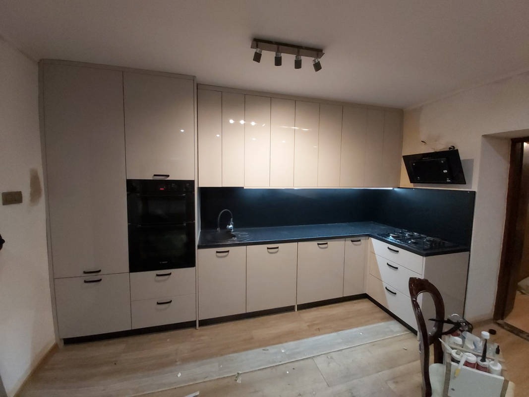 Beżowa kuchnia na wymiar z czarnym blatem i sprzętem AGD, oświetlona listwą LED pod szafkami wiszącymi. Widoczne detale wykończenia i minimalistyczne uchwyty.